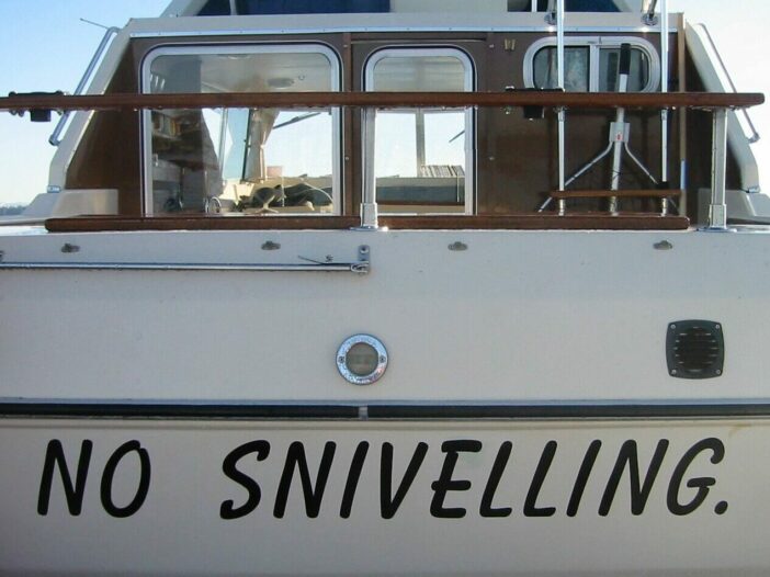 the name "No Sniveling" painted at the bow of a white sailboat with dark brown woodwork